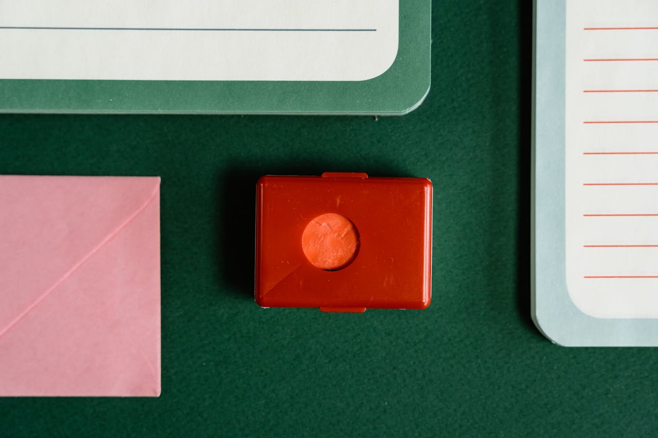 Flat lay of colorful stationery items including a red stamp, notebooks, and pink envelope on green background.